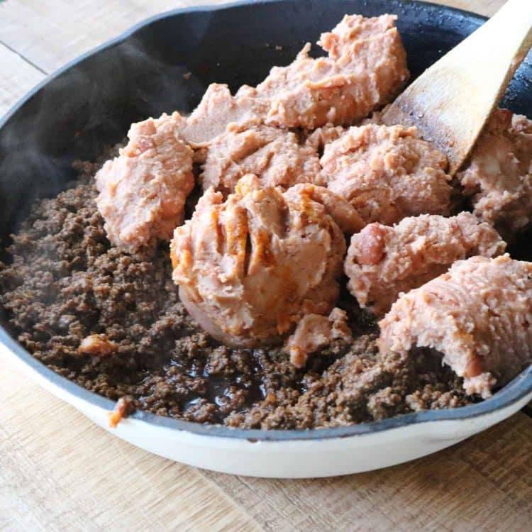 refried beans being cooked on ground beef with a wooden spoon in preparation for mexican pizza