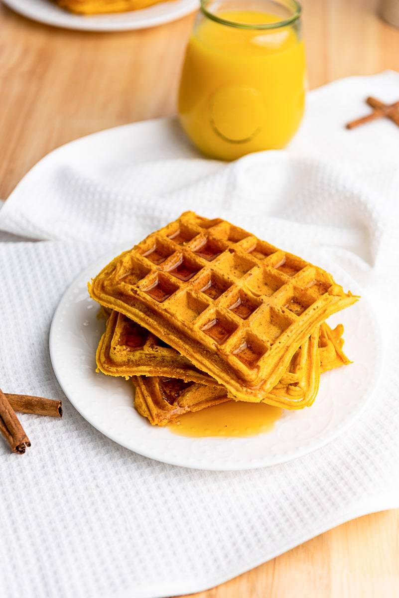 Easy pumpkin waffles stacked on a white plate with syrup, cinnamon sticks, and orange juice