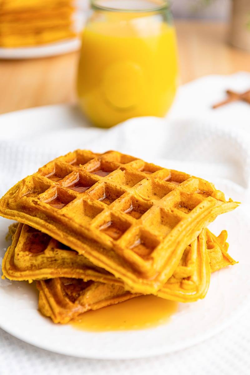 Stack of golden pumpkin waffles with syrup on a white plate, orange juice in the background