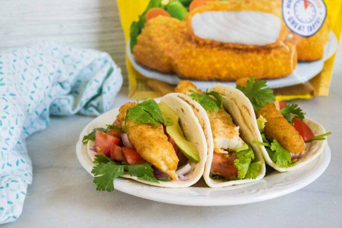 three fish tacos with pico sauce served on a white plate set in front of a bag of Gortons crunchy breaded fish fillets