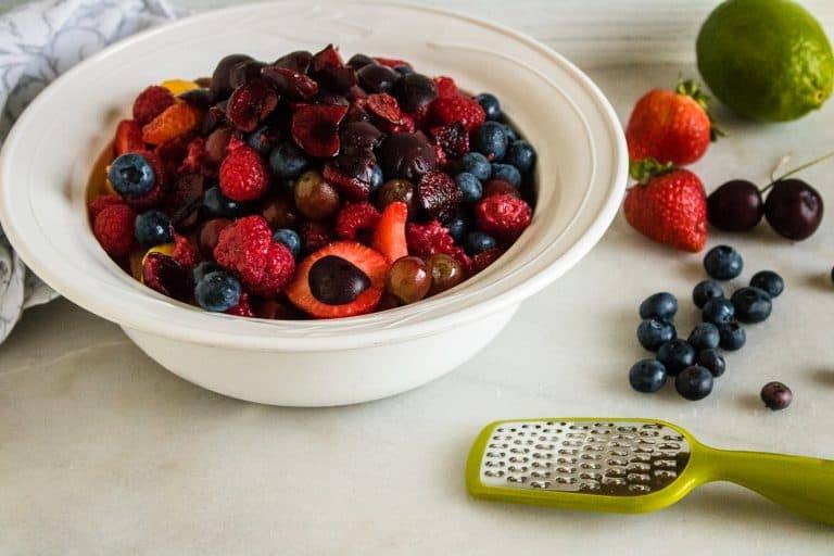 white ceramic bowl full of berries for fresh fruit salad with honey lime dressing with green zester and berries next to it