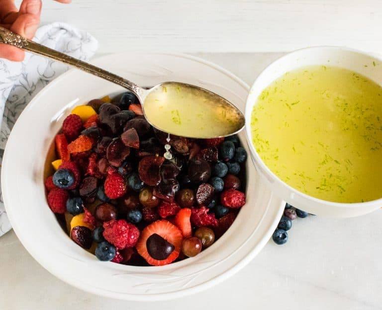 white bowl of mixed berries for fresh fruit salad with honey lime dressing with white bowl of honey lime dressing next to it and silver teaspoons drizzling dressing onto berries
