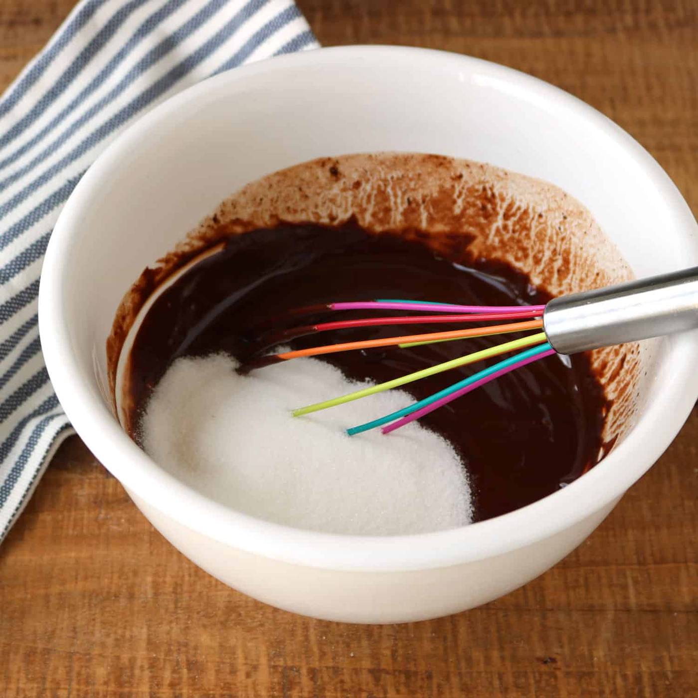 melted chocolate with sugar poured on top in a white bowl with a rainbow-colored whisk