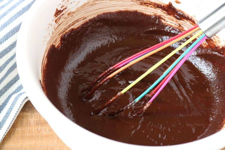 chocolate gluten free cake mixture in a white bowl with a rainbow-colored whisk