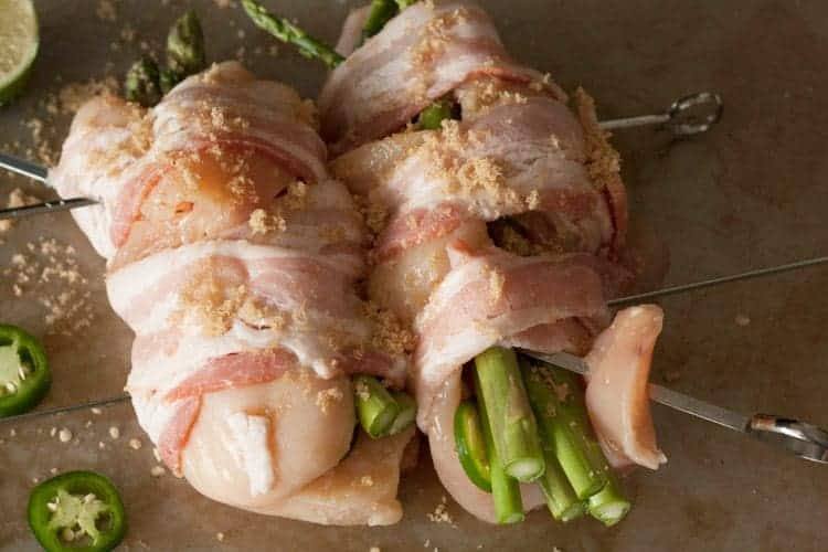 Two chicken breasts stuffed with asparagus and jalapeño, wrapped in bacon, sprinkled with brown sugar and threaded on metal skewers, ready for the grill