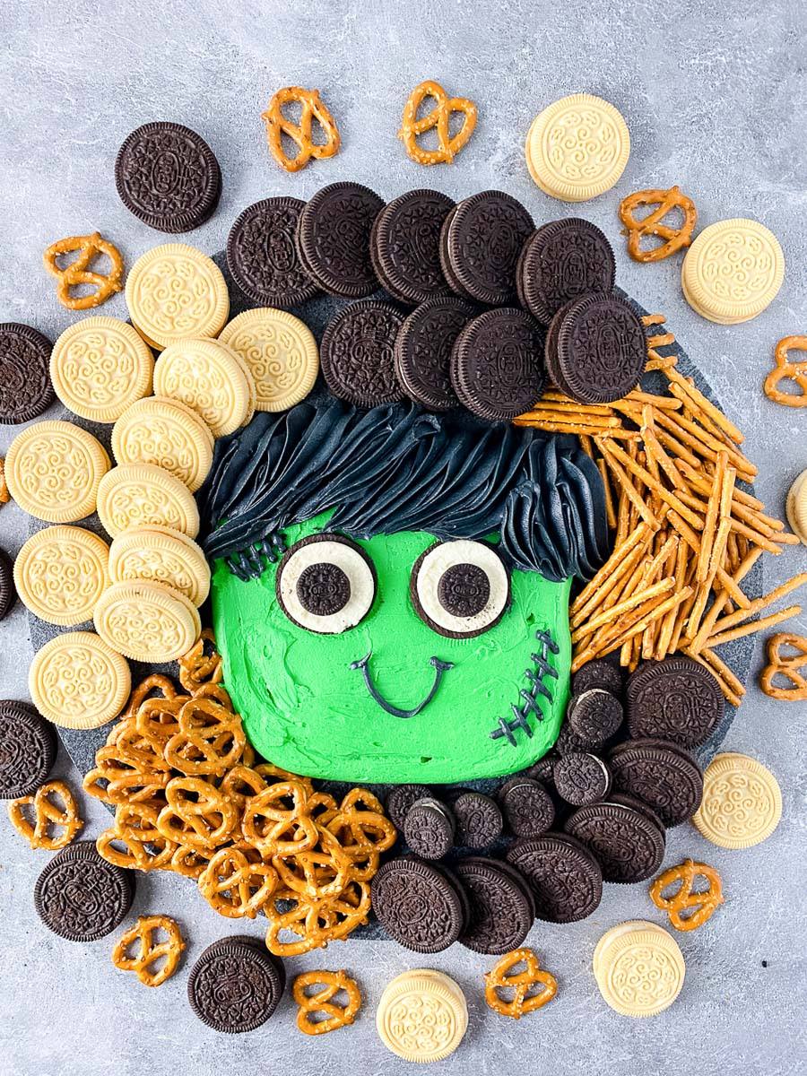 Halloween buttercream board with a green Frankenstein face, sandwich cookies and pretzels on a slate board