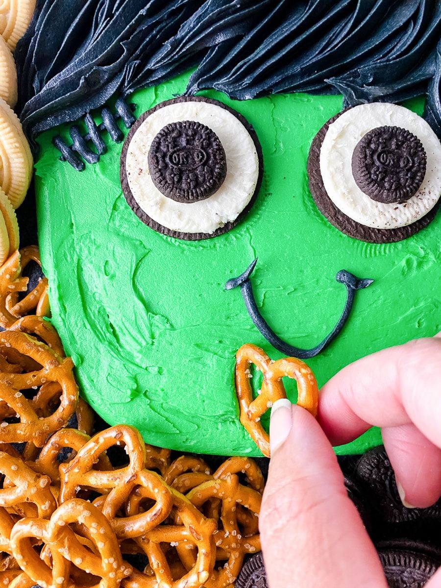 Close-up of a pretzel being dipped into the green frosting of a Frankenstein buttercream board