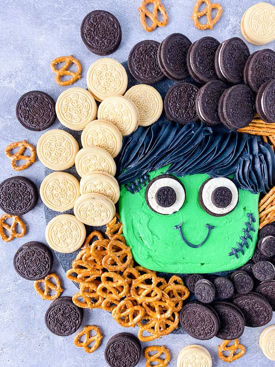 Frankenstein buttercream board surrounded by chocolate and vanilla sandwich cookies and mini pretzels