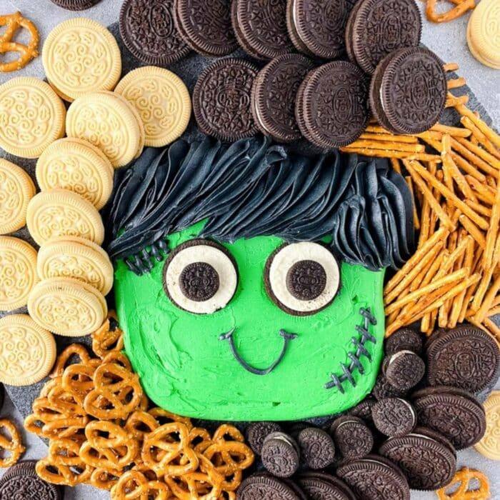 Halloween buttercream board with a green Frankenstein face surrounded by chocolate and vanilla sandwich cookies and pretzels