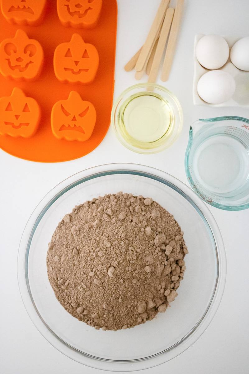 Ingredients for pumpkin cake pops including cocoa and flour mixture, oil, eggs, popsicle sticks and orange silicone pumpkin molds