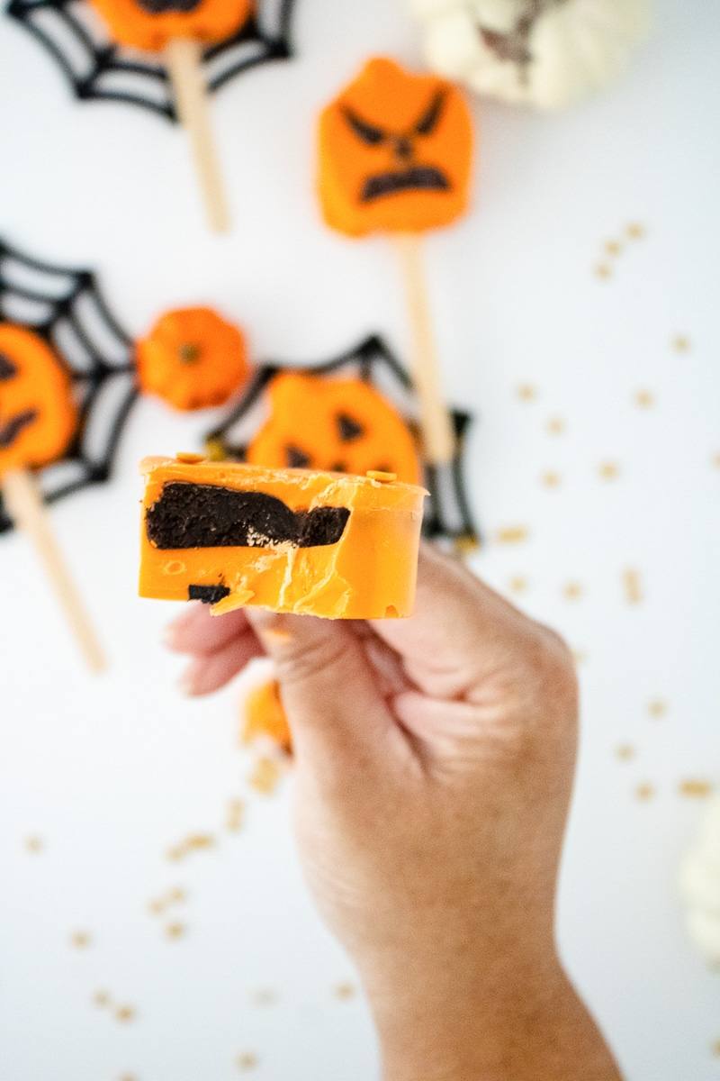 Hand holding a bitten pumpkin cake pop showing the chocolate cake center inside the orange candy shell