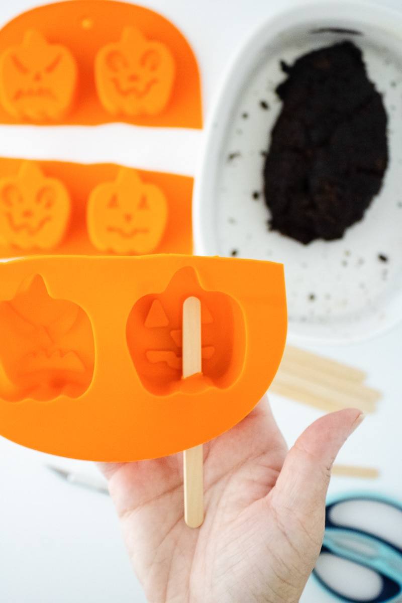 Hand holding an orange silicone pumpkin mold with a popsicle stick pushed through a slit in the bottom