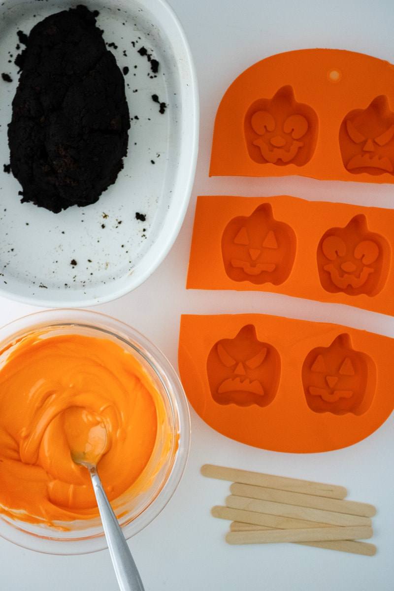 Bowl of melted orange candy melts next to chocolate cake crumbs and orange pumpkin molds