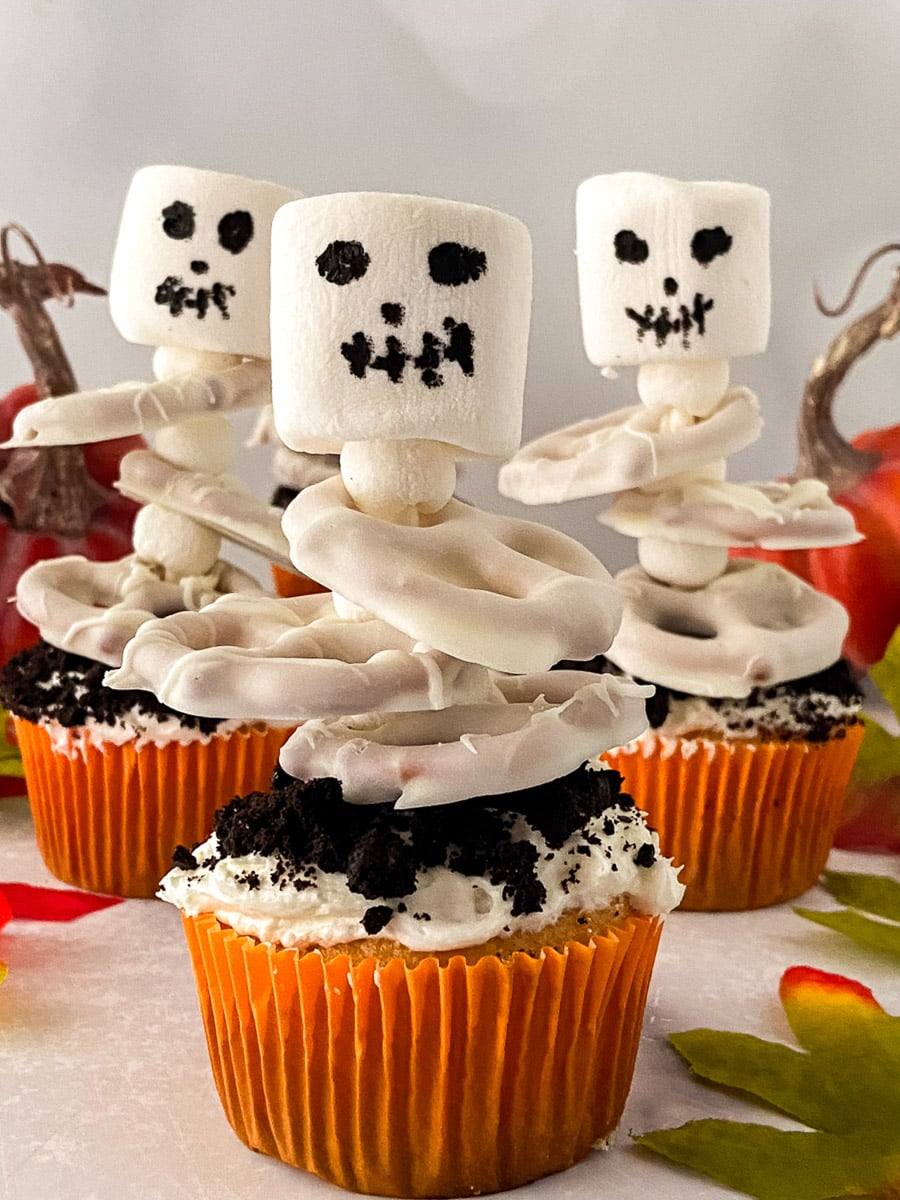 Halloween skeleton cupcakes with marshmallow skulls and white candy-coated pretzel ribcages, surrounded by mini pumpkins and fall leaves