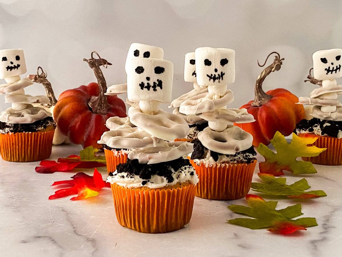 A row of skeleton cupcakes topped with crushed Oreos and marshmallow skeletons on a marble counter with mini pumpkins