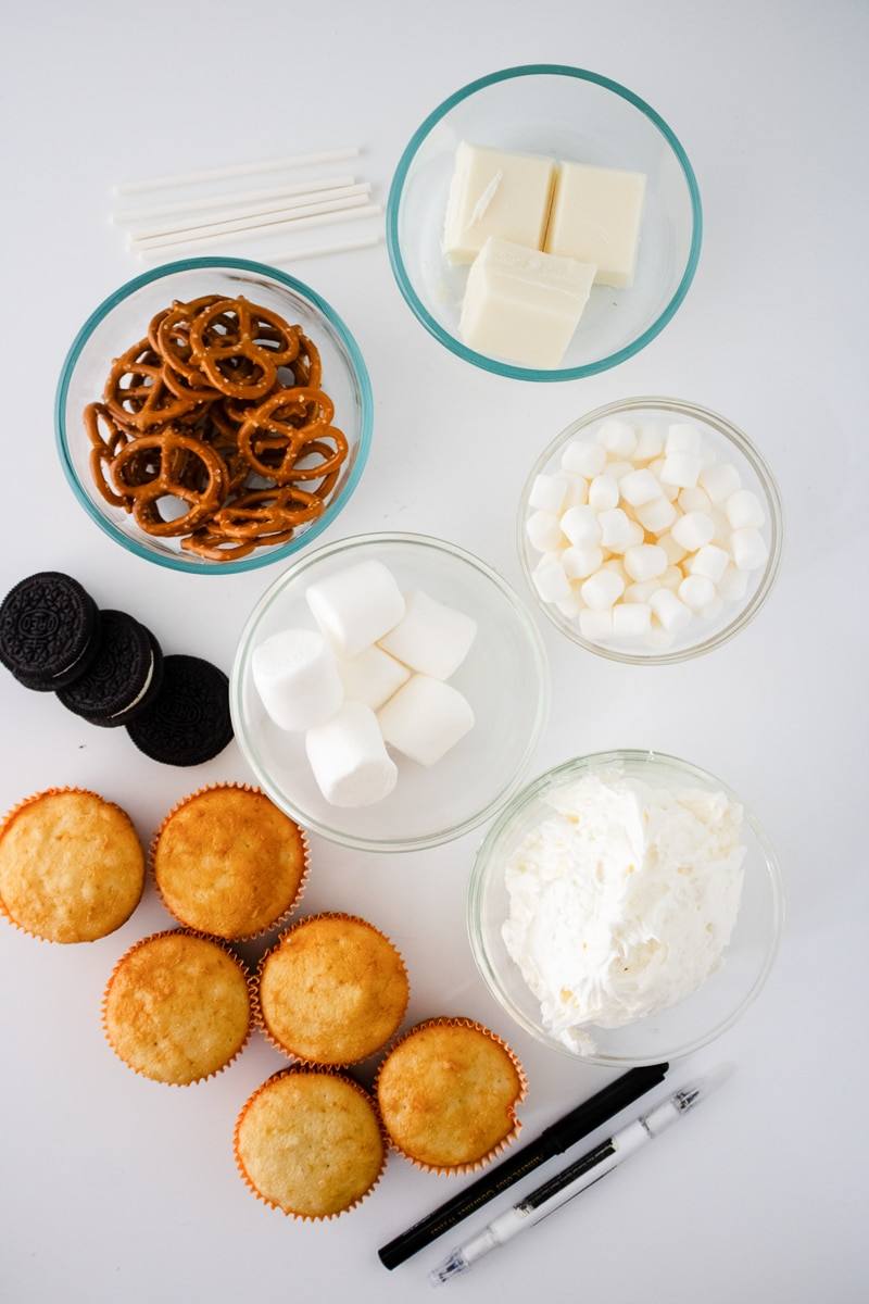 Ingredients for skeleton cupcakes laid out on a white counter: vanilla cupcakes, mini pretzels, white candy melts, large and mini marshmallows, Oreos, frosting, lollipop sticks and an edible marker
