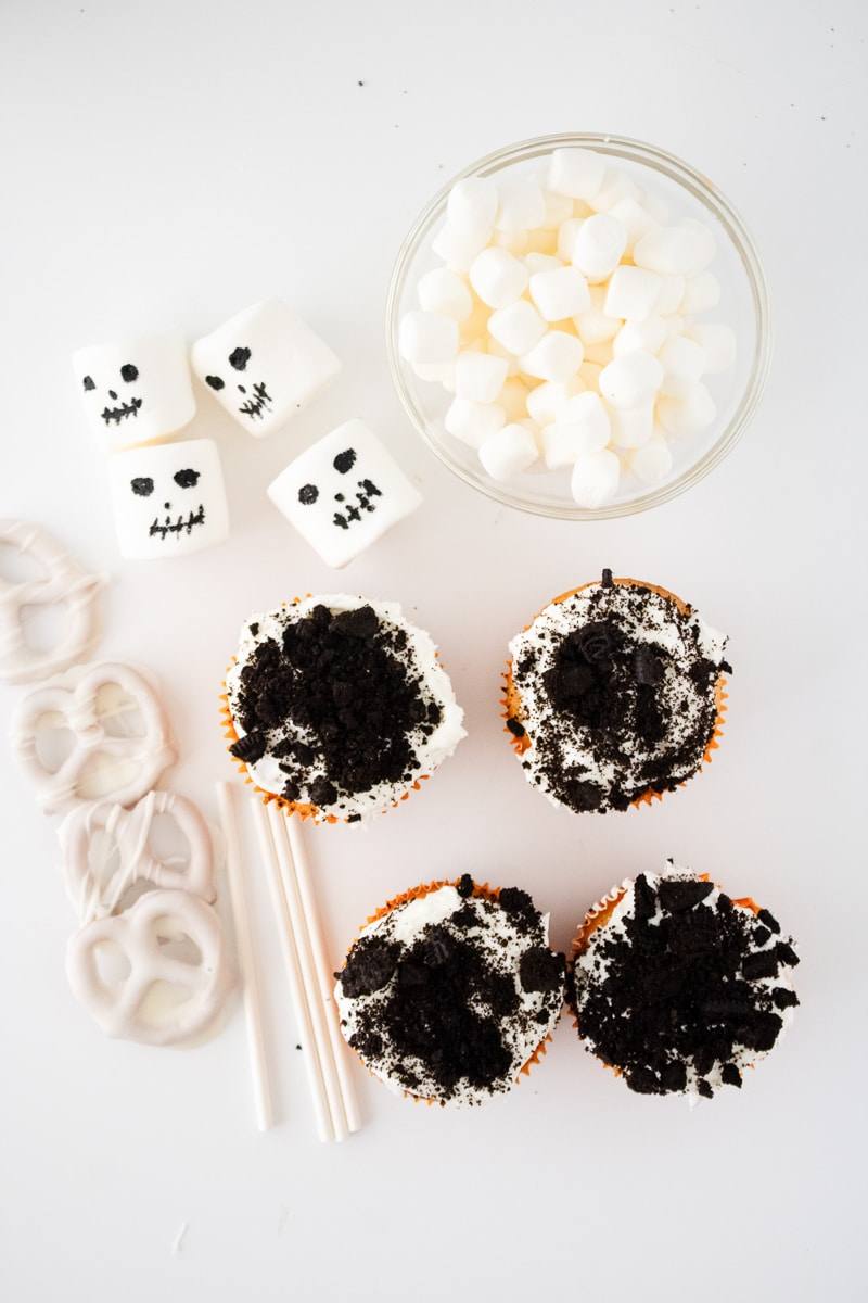 Cupcakes topped with frosting and crushed Oreo dirt next to candy-coated pretzels, mini marshmallows and marshmallow skeleton faces