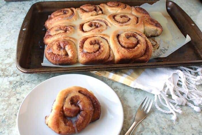 homemade cinnamon rolls sitting on a baking sheet in the background with a single cinnamon roll on a white plate in the foreground