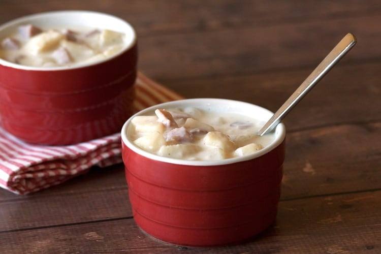 Two red ramekins of homemade ham and potato soup with a spoon on a wooden table