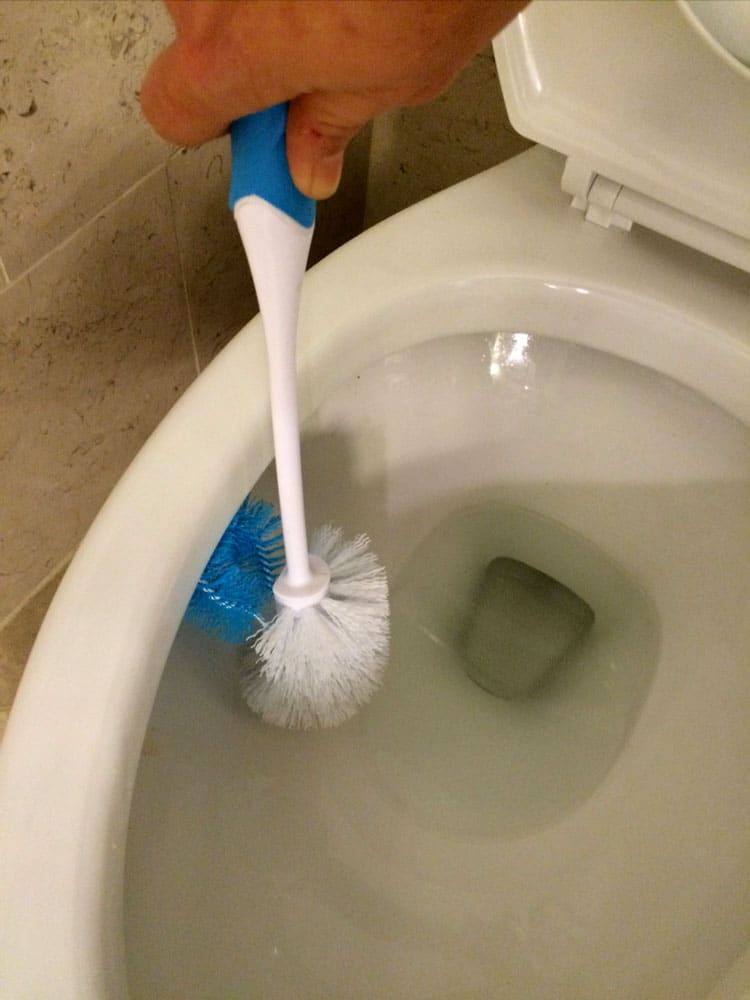 Gloved hand scrubbing the inside of a toilet bowl with a white toilet brush that has a blue under-rim brush