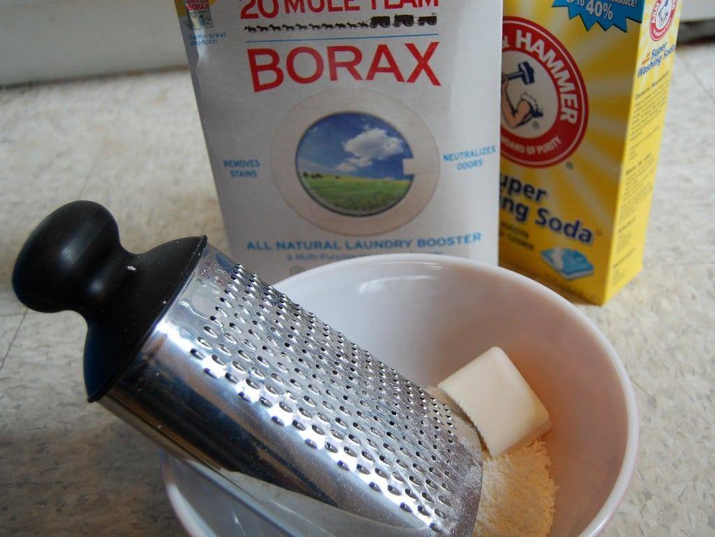 Borax, washing soda and a bar of soap being grated into a white bowl to make homemade laundry detergent