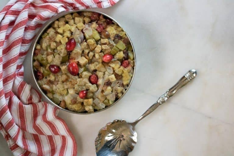 The completed Instant Pot Cranberry Pecan Stuffing in a metal pan sitting next to a silver serving spoon and red striped dish towel