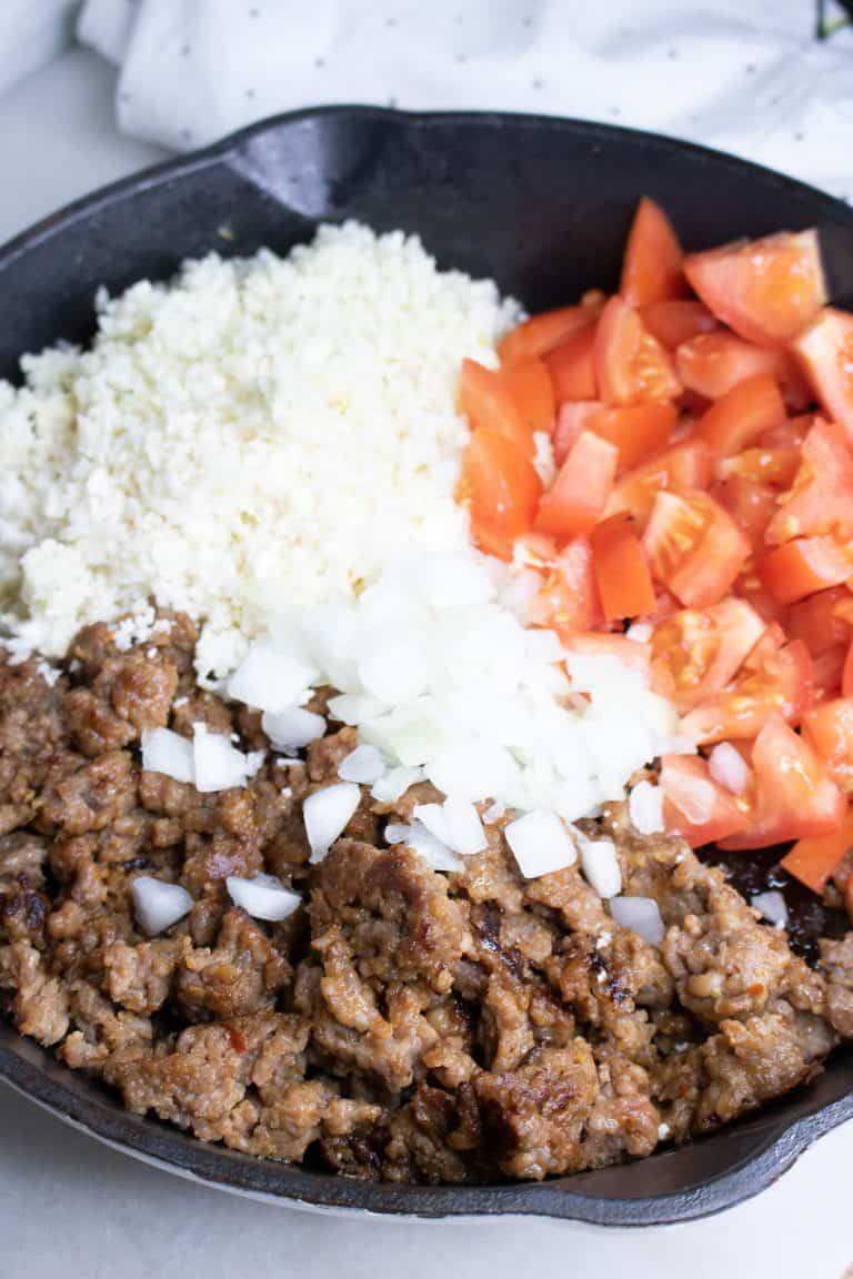 Cauliflower rice, cooked Italian sausage, chopped onions and chopped tomatoes in an iron skillet