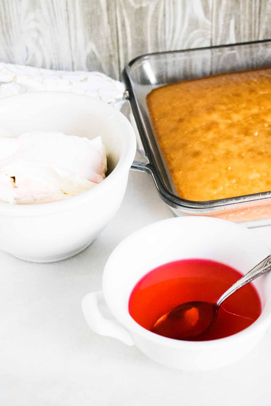 baked white cake in a sheet pan next to a bowl of Jolly Rancher Watermelon Gelatin liquid and a larger bowl of creams cheese mixture