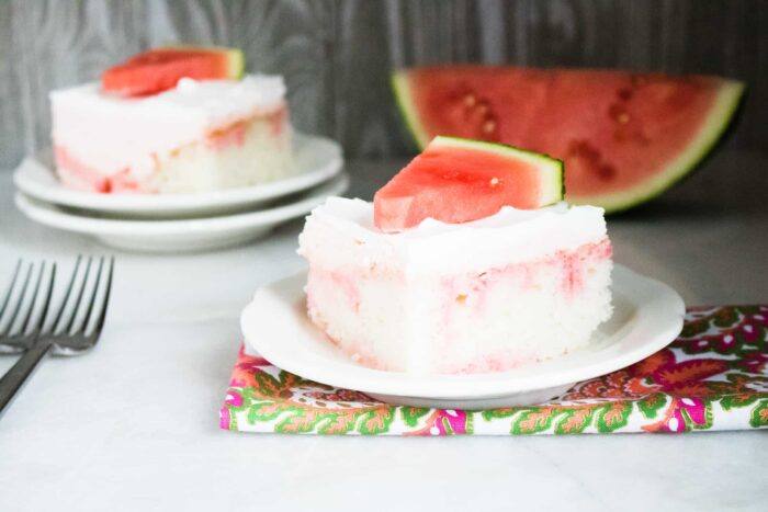 A slice of Watermelon Jolly Rancher Poke Cake served on a white dessert plate on top of a floral print cloth napkin