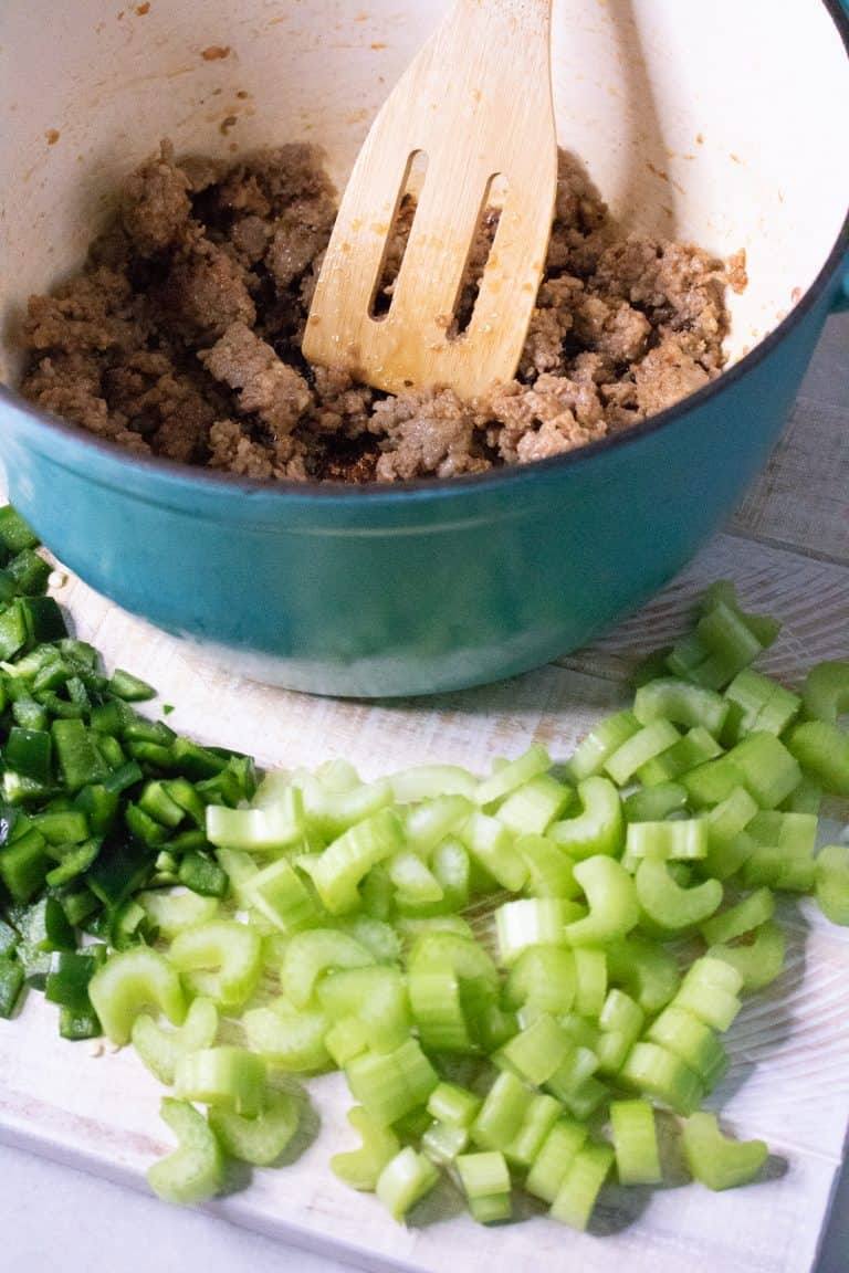 Diced celery and Poblano pepper on a cutting board next to browned sausage in a Dutch oven