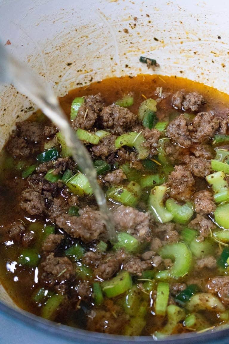 Chicken stock being poured into a pot of browned sausage and vegetables