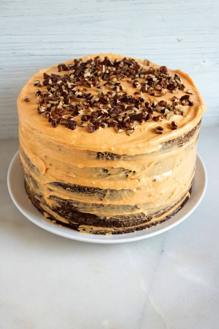 Layered chocolate cake with pumpkin cream cheese frosting topped with chopped pecans on a white plate