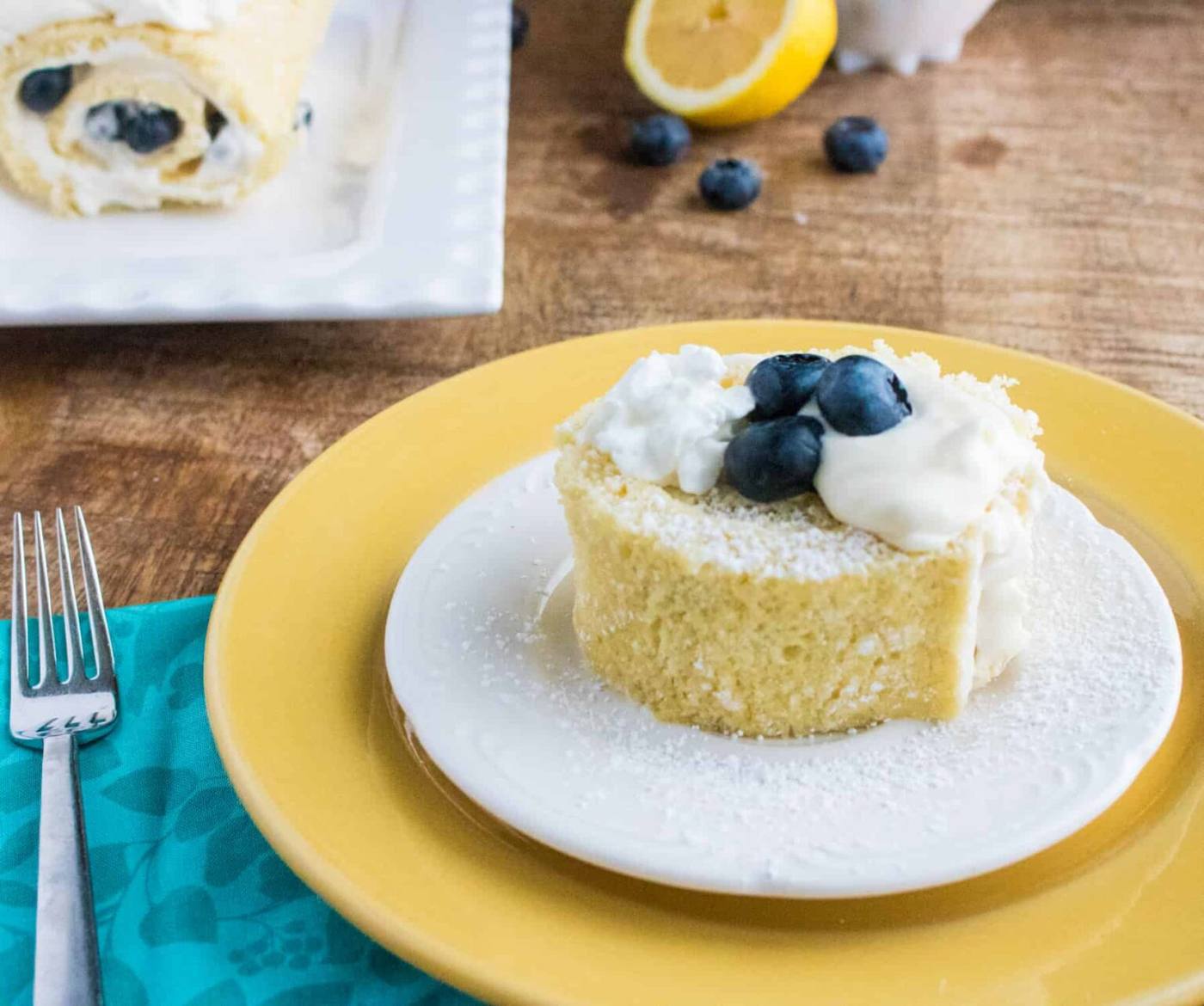 A slice of a Lemon Blueberry Cake Roll on a white saucer on top of a yellow plate next to a fork on a teal napkin