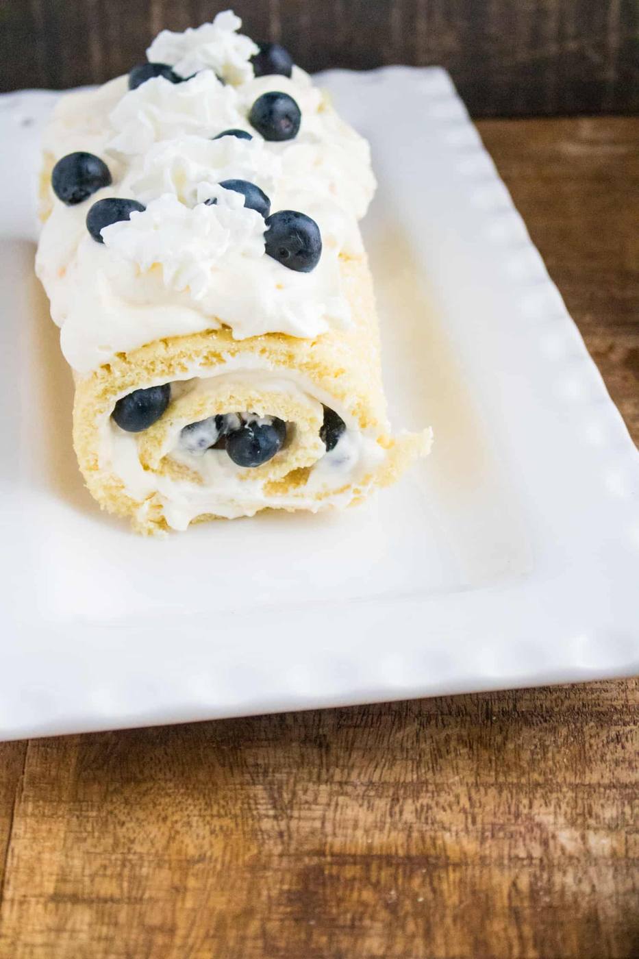 A Lemon Blueberry Cake Roll topped with whipped cream and sprinkled with blueberries on a white plate