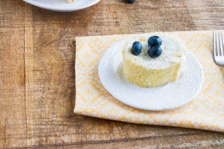 a slice of Lemon Blueberry Cake Roll topped with three blueberries on a white saucer on top of a peach colored printed napkin