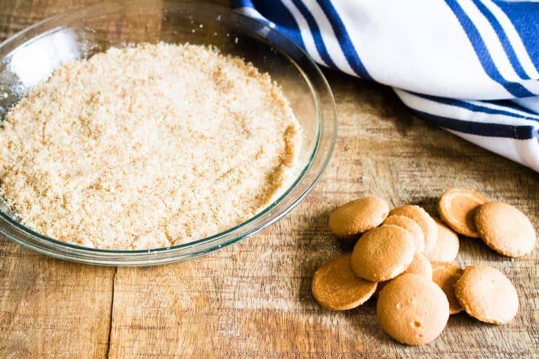 crushed vanilla wafers for no bake tropical cream pie in clear pie plate on the left with a pile of Nilla wafers on the right and a blue and white striped dish towel in background