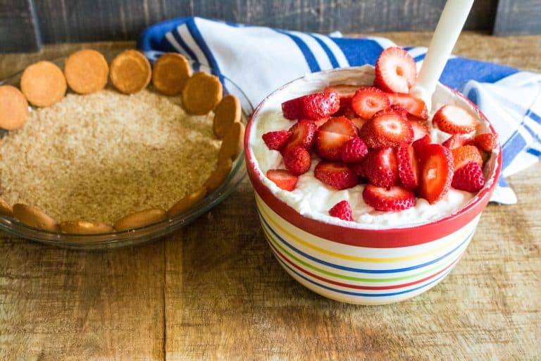 clear pie plate with crushed Nilla wafers for no bake tropical cream pie on the bottom and whole Nilla wafers lining the edge. To the right is a red, yellow, blue, green and white striped ceramic bowl with pie filling in it with sliced strawberries on top and white spoon at the side. There is a blue and white dish towel in background