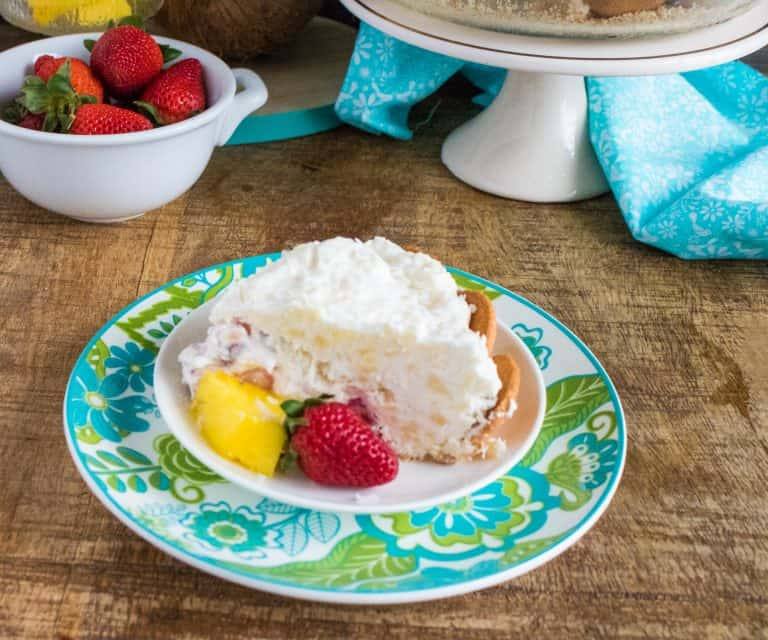 piece of no bake tropical cream pie on a small with dish sitting on a blue and green liner plate with white bowl of strawberries in background next to a white cake stand holding the pie plate of no bake tropical cream pie