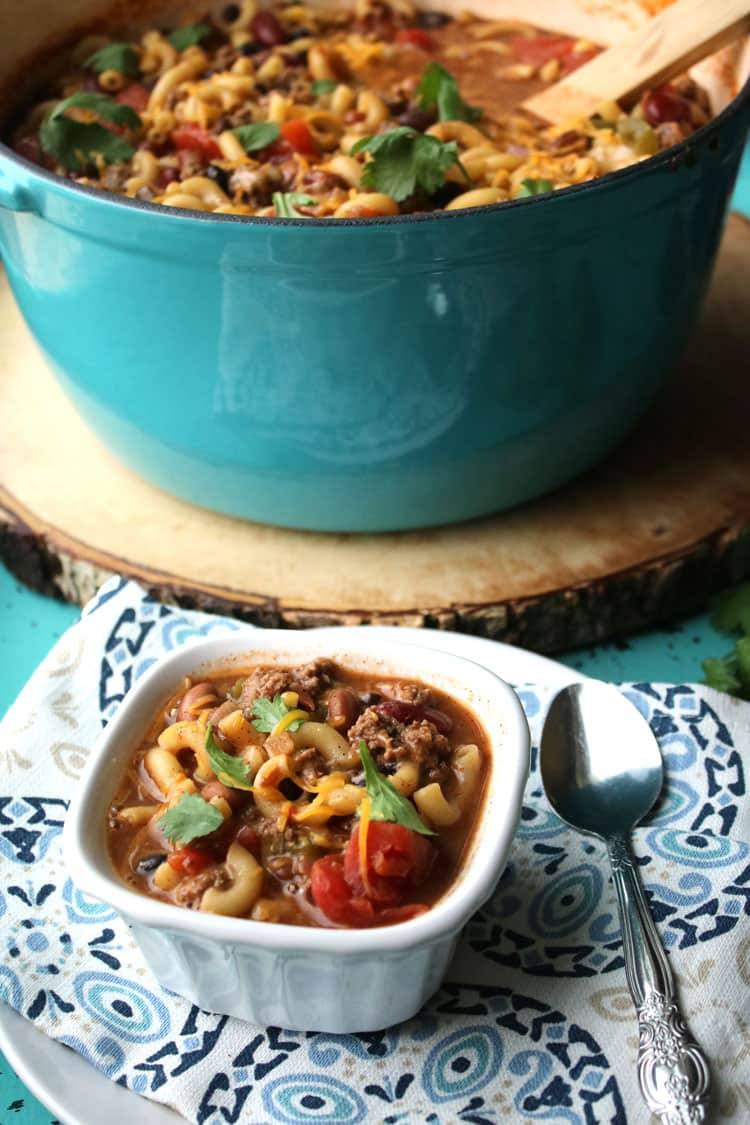 A white bowl of chili mac and cheese with cilantro in front of a blue pot of chili mac