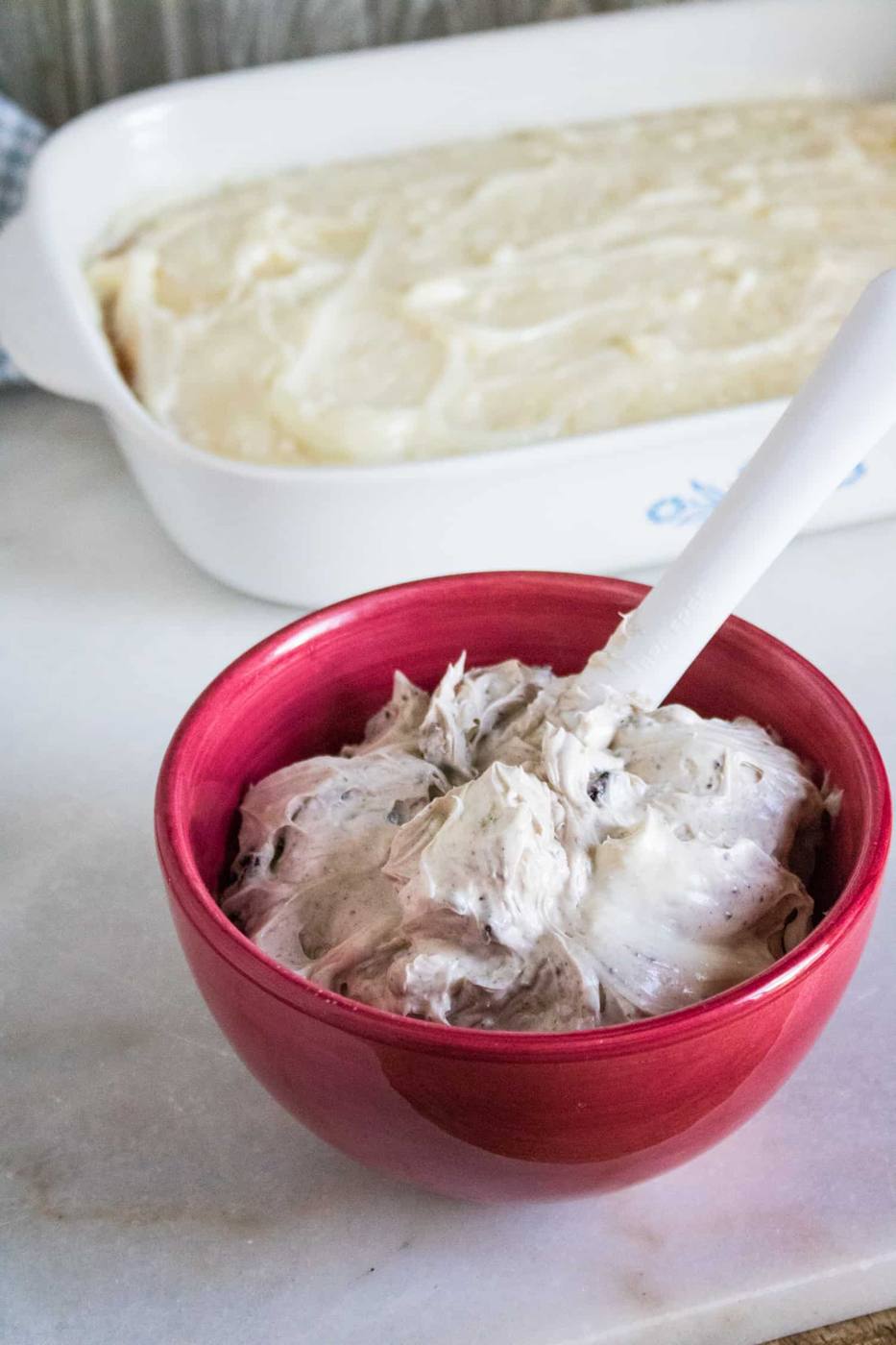 background has white 13x9 corning ware pan with cake for oreo poke cake spread in it. foreground has red mixing bowl with cream cheese oreo topping in it being stirred by a white spoon