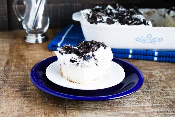 A slice of Oreo Poke Cake served on a white and blue dessert plate and sitting in front of the rest of the Oreo Poke Cake in a white corning ware sheet pan