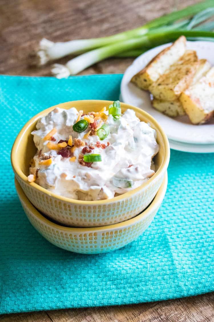 baked potato dip in small yellow bowl stacked on top of another yellow bowl on a table with a blue cloth