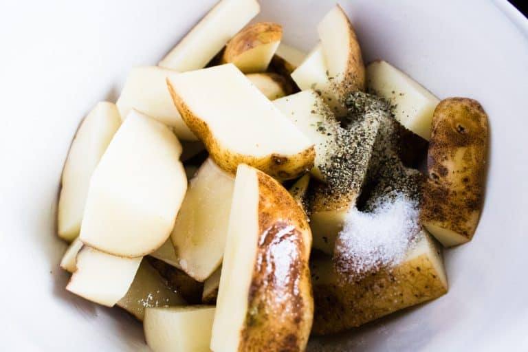 close up view of sliced chunks of potatoes in a white mixing bowl with Italian seasoning sprinkled on top
