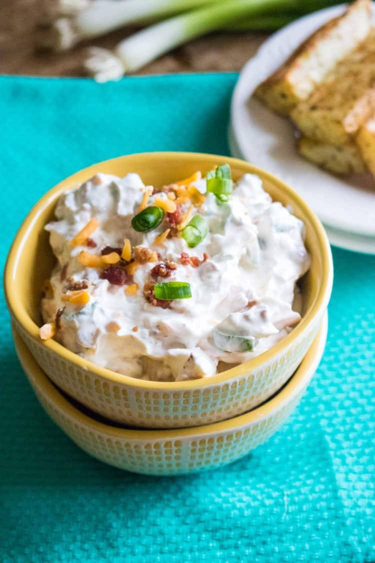 baked potato dip in small yellow bowl stacked on top of another yellow bowl on a table with a blue cloth