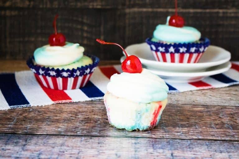 one unwrapped patriotic poke cupcake in front of two other patriotic poke cupcakes sitting on top of red white and blue striped table runner