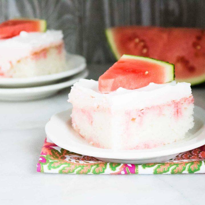 A slice of Watermelon Jolly Rancher Poke Cake served on a white dessert plate on top of a floral print cloth napkin