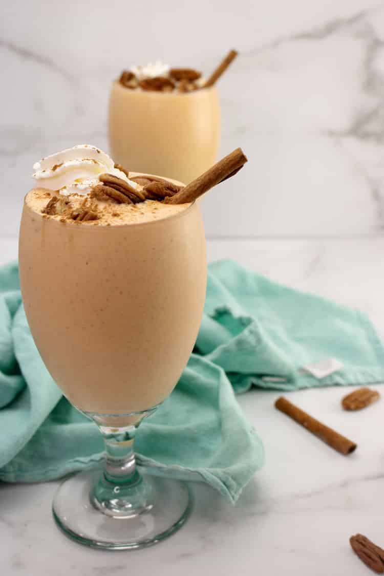 Two pumpkin cheesecake milkshakes in footed glasses topped with whipped cream, pecans and cinnamon sticks