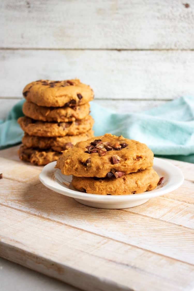 Pumpkin chocolate chip cookies topped with chopped pecans on a white plate in front of a tall cookie stack