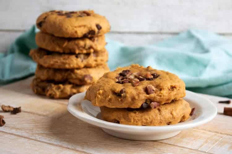 A stack of pumpkin chocolate chip cookies behind a plate of two cookies on a rustic wooden board