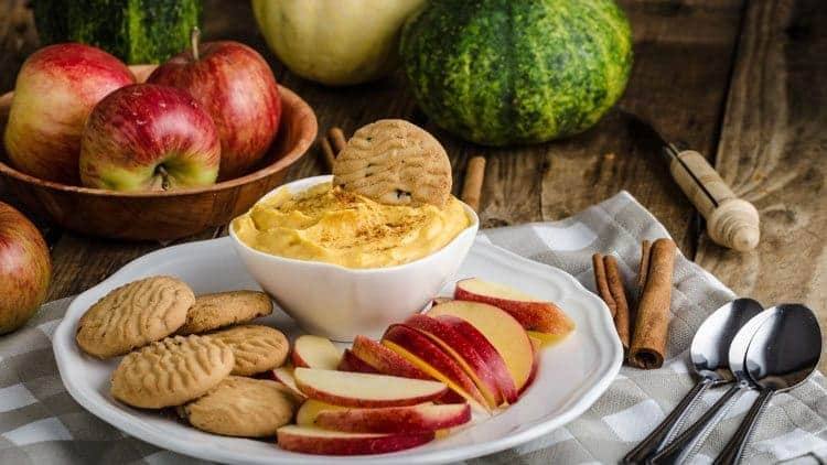 Bowl of pumpkin cream cheese filling topped with cinnamon, served with apple slices and cookies on a white plate